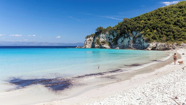 Een strand met helder turquoise water, witte kiezelstenen en een rotsklif met begroeiing.