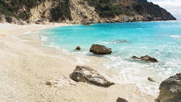 A beach with clear turquoise water, rocky outcrops, and a forested hillside in the background.
