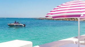 A beachside view with a boat on the water, a white umbrella, and a table with chairs.
