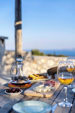 Une table dressée avec une carafe en verre, du fromage, des olives et de la charcuterie, accompagnée d’un verre de vin, et une vue sur la mer en arrière-plan.