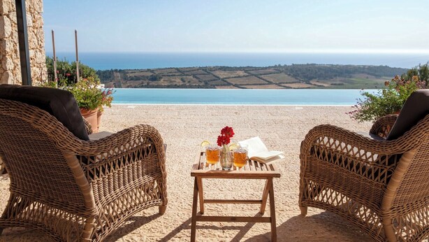 Un juego de sillón y mesita de mimbre sobre una playa arenosa, con vista al mar y colinas lejanas.
