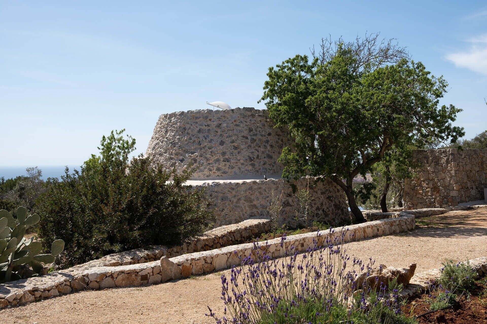 Uma fortificação de pedra com vista ampla para o mar, rodeada por cactos e plantas de lavanda.