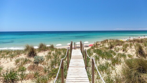 A wooden boardwalk leading to a beach with clear turquoise water and a clear sky.