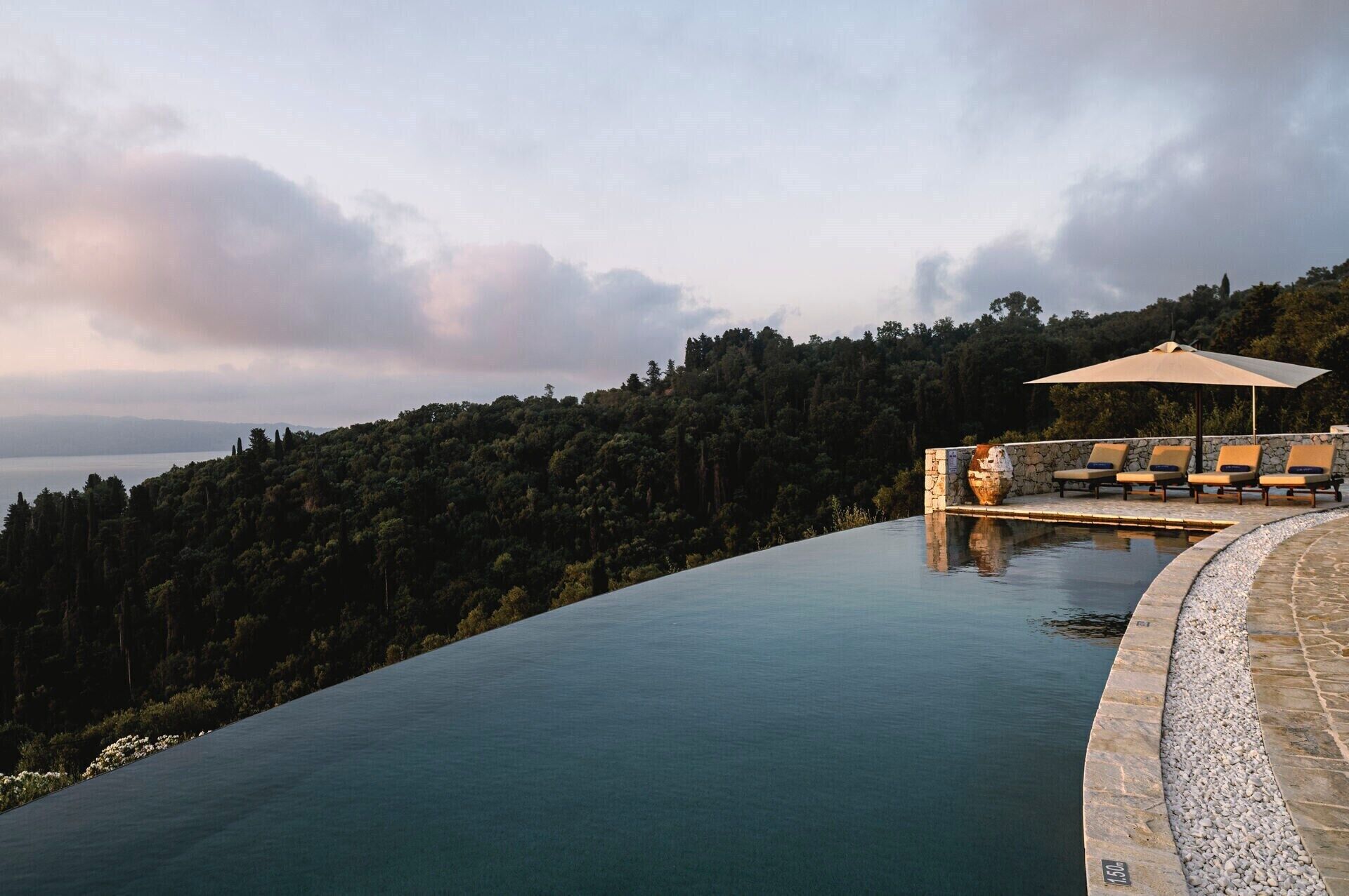 A infinity pool with a stone edge, a lounge area with chairs and an umbrella, and a forested hillside in the background.
