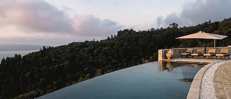 En infinitypool med en kant av sten, ett område med solstolar och ett parasoll, och en skogbeklädd sluttning i bakgrunden.