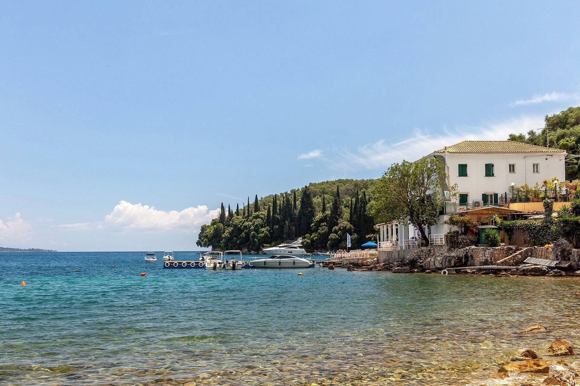 A coastal scene with a white building with green shutters, a rocky shoreline, and boats in the water.