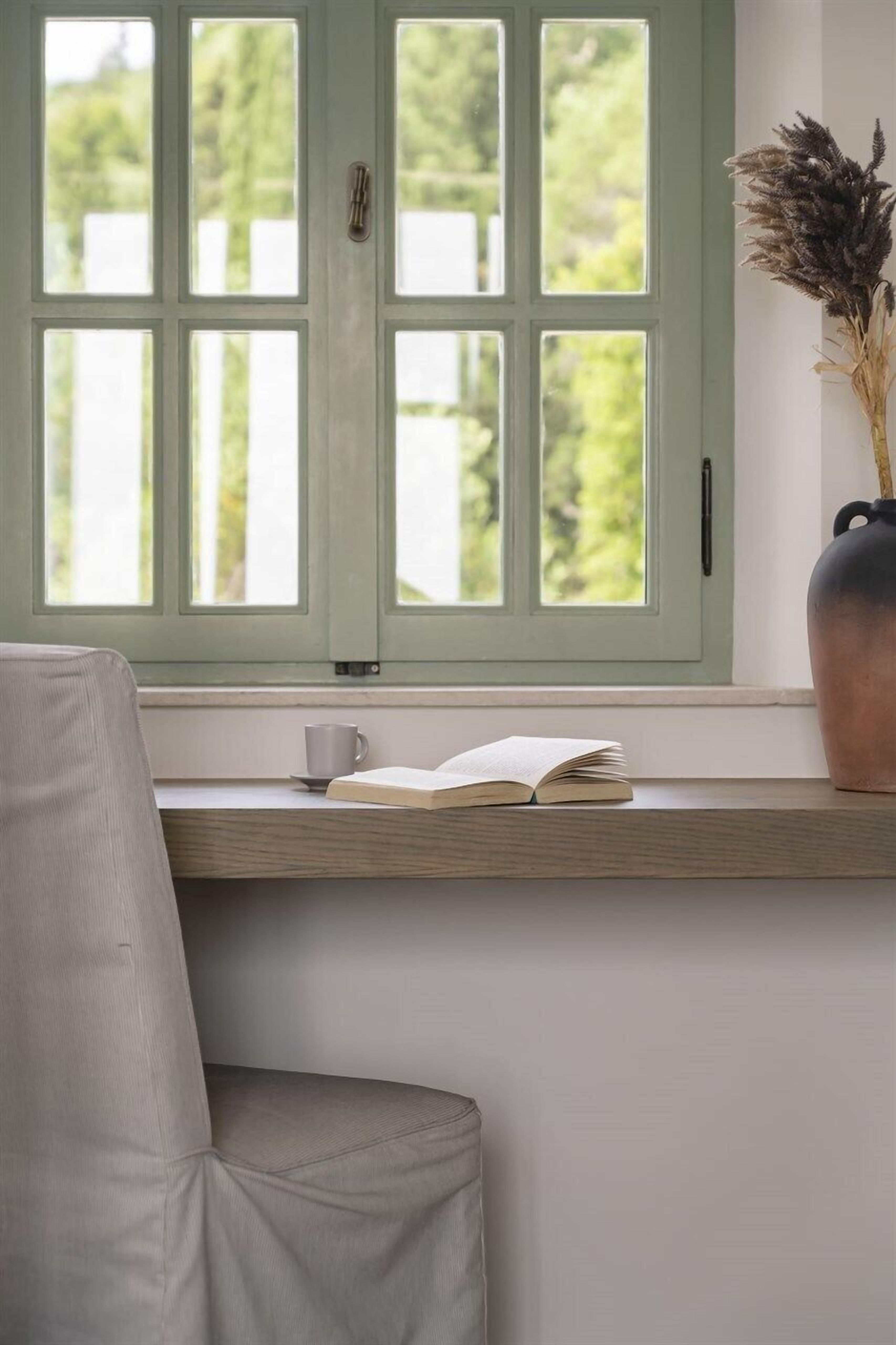 A room with a window, a chair, a table, and a vase with dried plants.