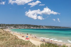 Une plage avec une eau turquoise et limpide, des gens qui se prélassent au soleil, et une ville en arrière-plan.