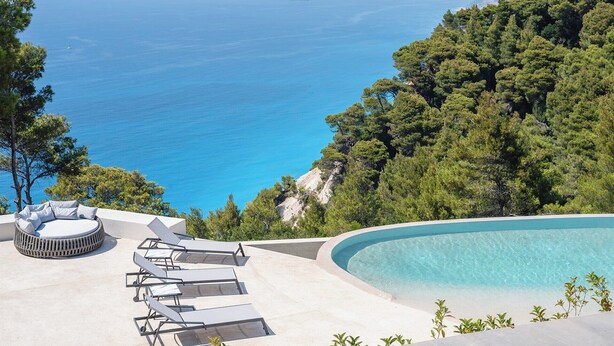 A poolside area with lounge chairs and a circular seating area overlooking a clear blue sea and dense green forest. - Noiya - Eight Bedroom Villa, Sleeps 16 (Ponti Agiou Petrou)
