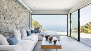 A modern living room with a stone wall, a sofa, a coffee table with bowls, and a large window overlooking a scenic view. - Noiya - Eight Bedroom Villa, Sleeps 16 (Ponti Agiou Petrou)