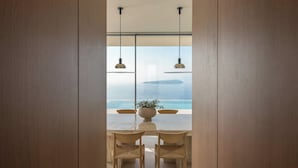 A dining area with a table and chairs, a potted plant, and a view of the sea through large windows. - Oikos Nostos - Three Bedroom Villa, Sleeps 6 (Lefkada City)