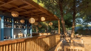 Een buitengebied met een bar, houten planken vol flessen, hangende rieten lampen en houten tafels en stoelen.