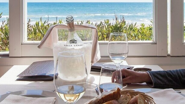 Tafel gedekt voor twee personen, met een fles Villa Sandi wijn, twee glazen en een mand met brood, met uitzicht op een strand.