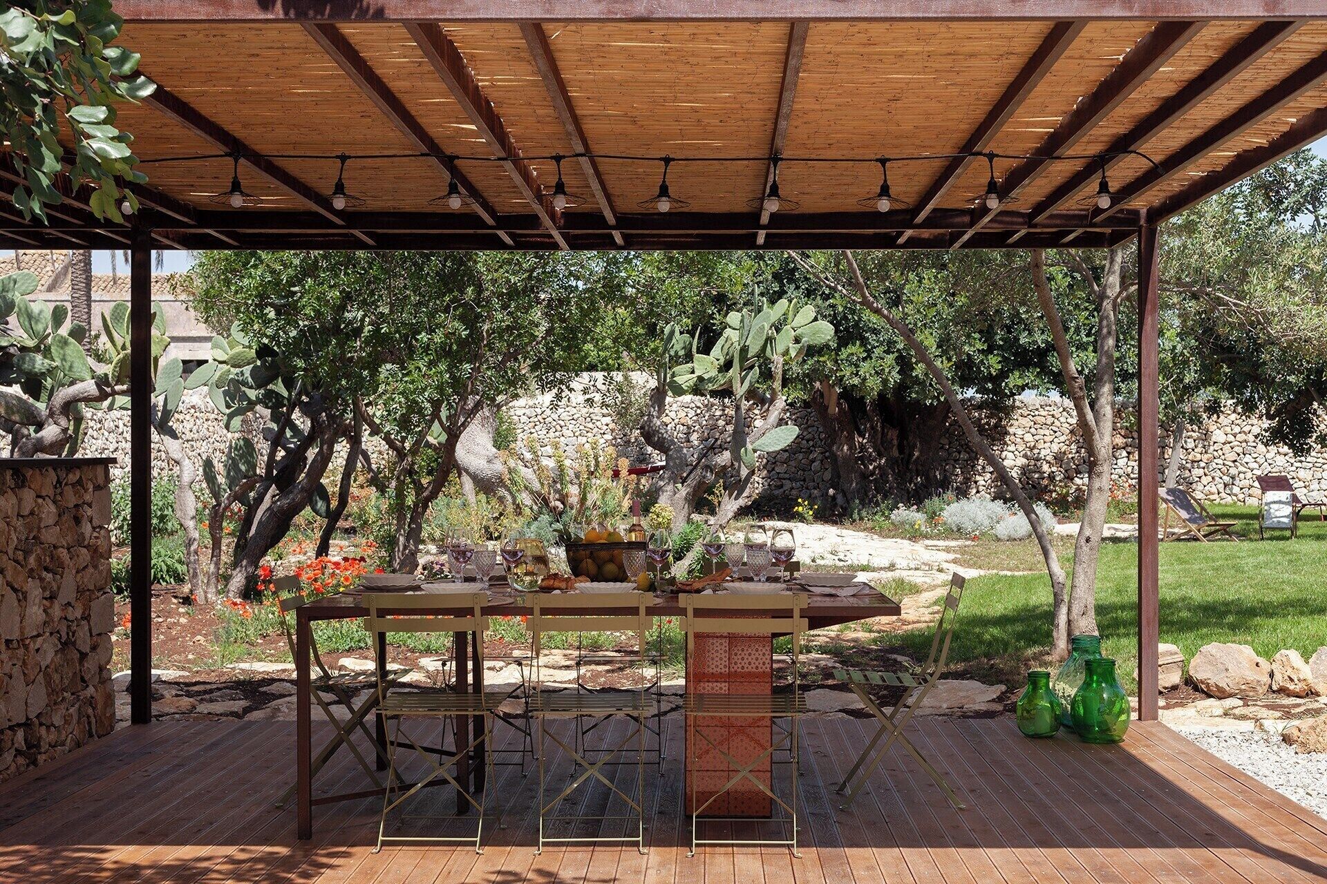 An outdoor patio with a wooden pergola, a table set with wine glasses, and a rustic stone wall in the background.