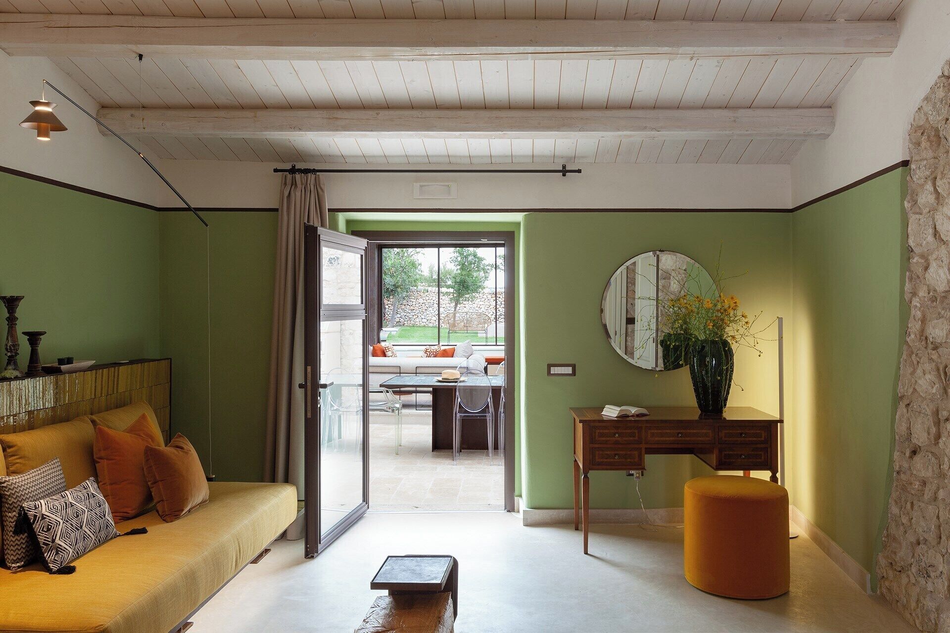 Een kamer met een bank, een ronde tafel en een stoel. Er is een raam met gordijnen, een spiegel aan de muur en een vaas met bloemen.