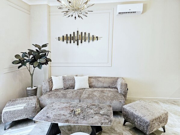 A living room with a sofa, two ottomans, a coffee table, a potted plant, a chandelier, and a wall-mounted air conditioner. - DiAngelo Guesthouse (Yerevan)