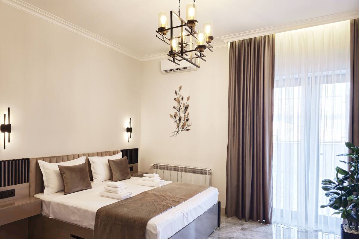 A modern hotel room with a large bed, a chandelier, a radiator, and a potted plant.