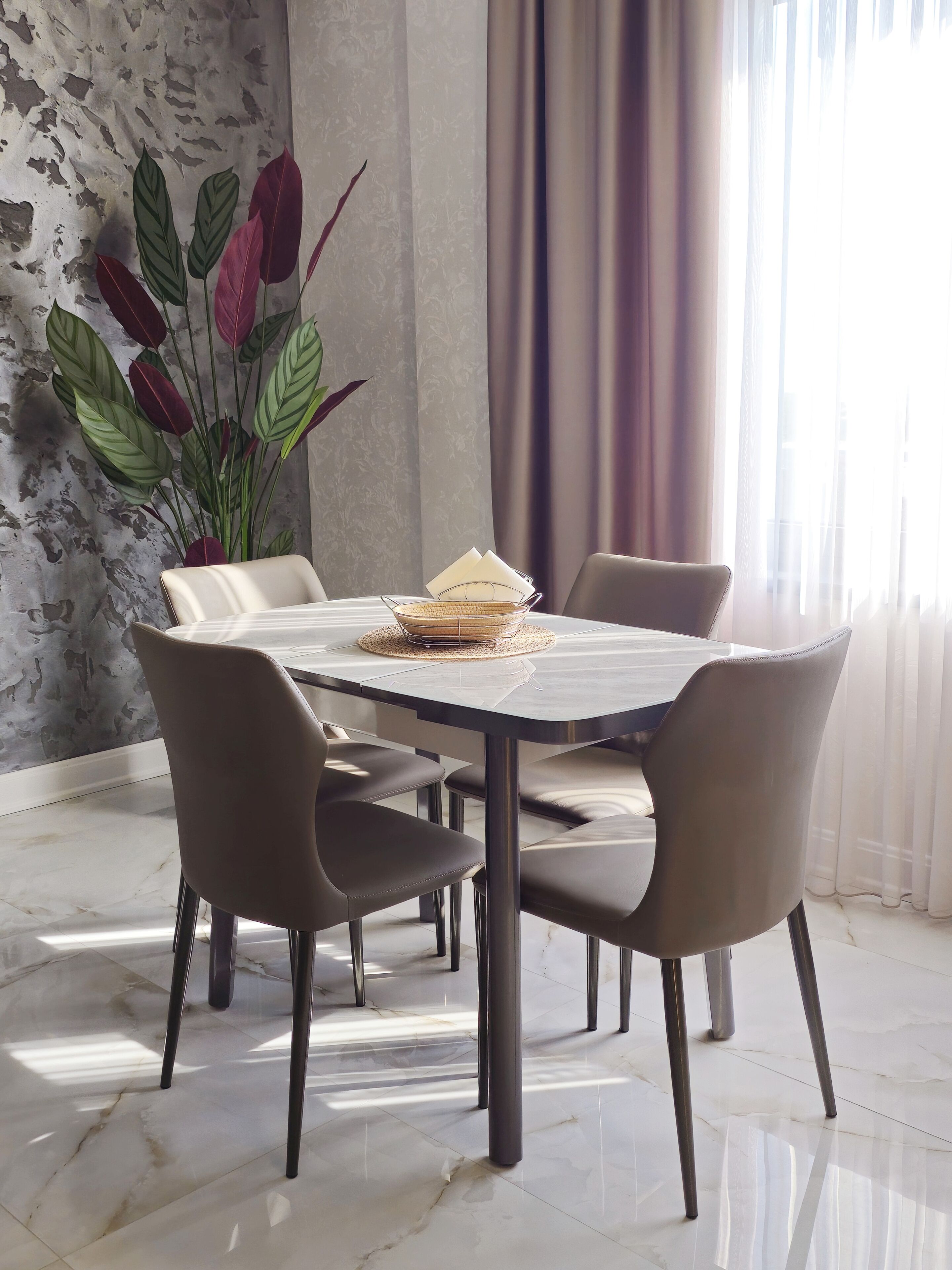 A dining area with a round table, six chairs, a vase with red and green leaves, and a window with curtains.
