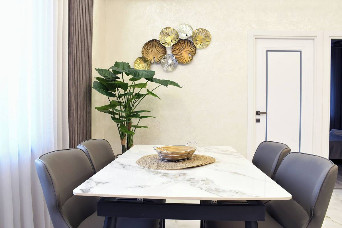 A dining table with a white marble top, surrounded by four grey upholstered chairs. A woven placemat is placed on the table, and a woven basket is positioned in the center.