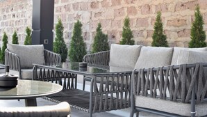 Un área de estar al aire libre con un sofá y sillones, una mesa de vidrio y un muro de ladrillo al fondo.