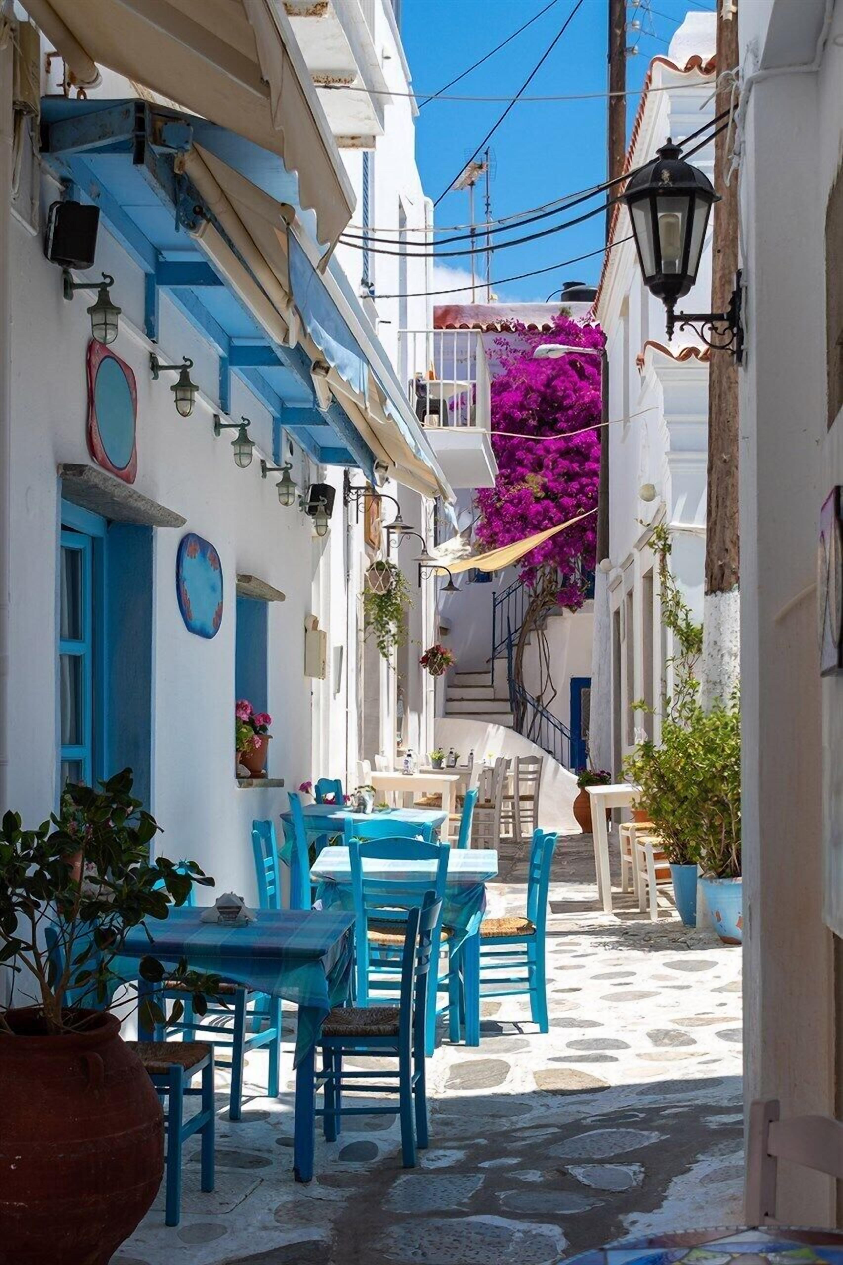 A narrow street with white buildings, blue doors, and tables with blue chairs.