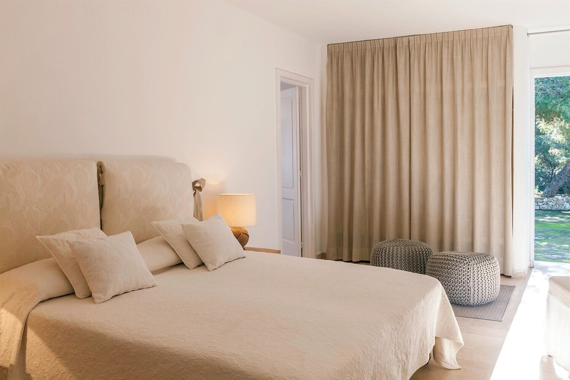 Ein geräumiges Schlafzimmer mit einem großen Bett, einer Nachttischlampe, einem Fenster mit Vorhängen und Blick auf Grünfläche.