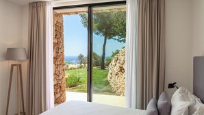 A bedroom with a large bed, a lamp, and a view of a stone wall and greenery outside.