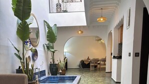 A spacious indoor area with a pool, large potted plants, a dining area, and a high ceiling with a decorative light fixture. - Riad Marrakech Médina renovated, 5 air-conditioned rooms, with swimming pool (MARRAKECH)