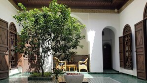 A courtyard with a large tree, a stone fountain, and traditional wooden doors. - Riad Marrakech Médina renovated, 5 air-conditioned rooms, with swimming pool (MARRAKECH)