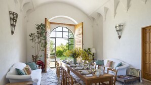 Un comedor con una larga mesa de madera dispuesta para seis personas, rodeado de sofás blancos y sillas de madera. Un arco abierto conduce a un jardín con flores amarillas.