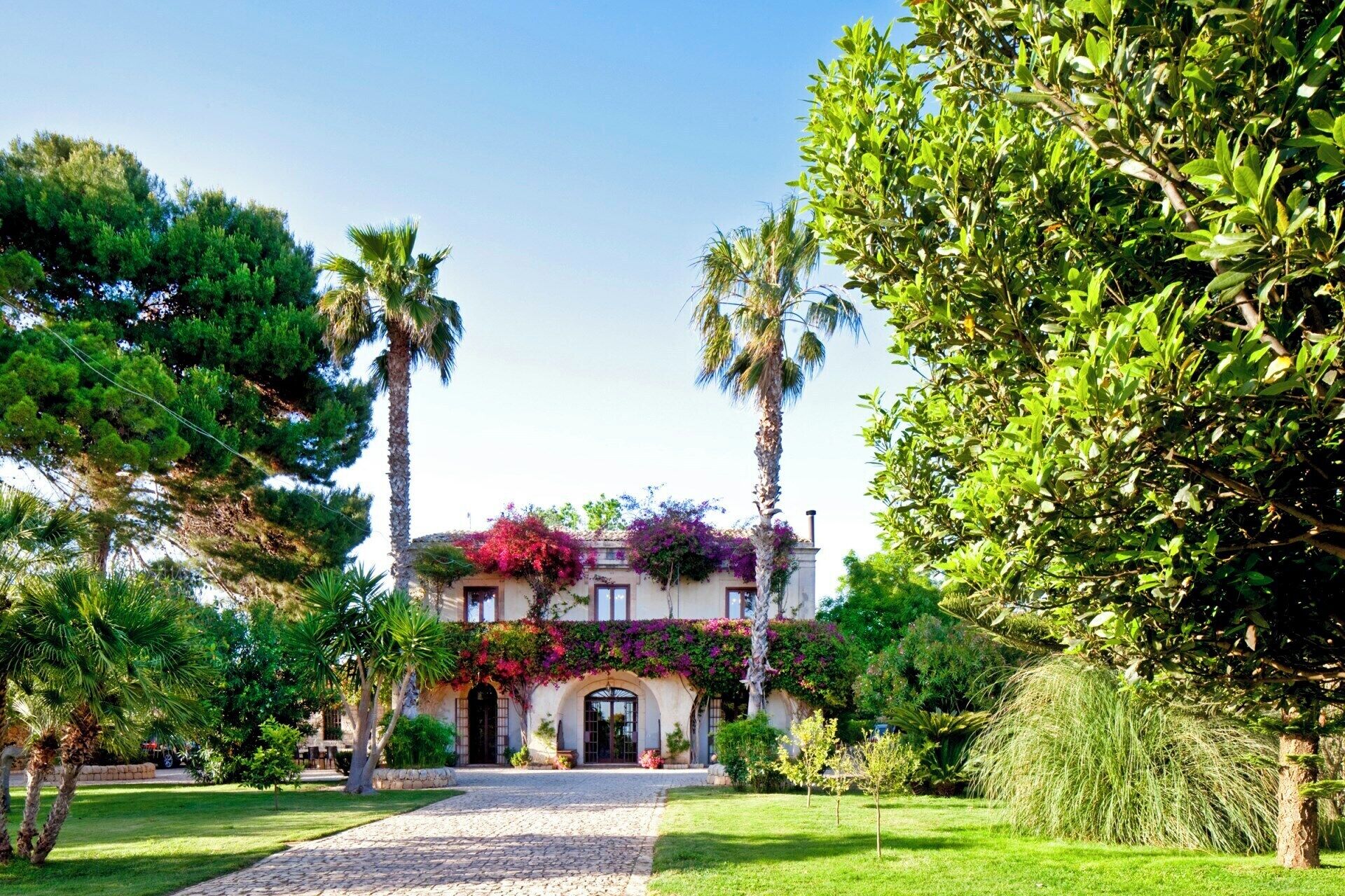 Eine Villa im mediterranen Stil mit Kopfsteinpflasterauffahrt, umgeben von üppiger Vegetation und Palmen.