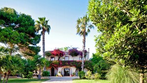 A Mediterranean-style villa with a cobblestone driveway, surrounded by lush greenery and palm trees.