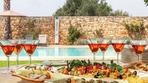 A dining table with a variety of dishes and drinks, a stone wall, and a pool in the background.