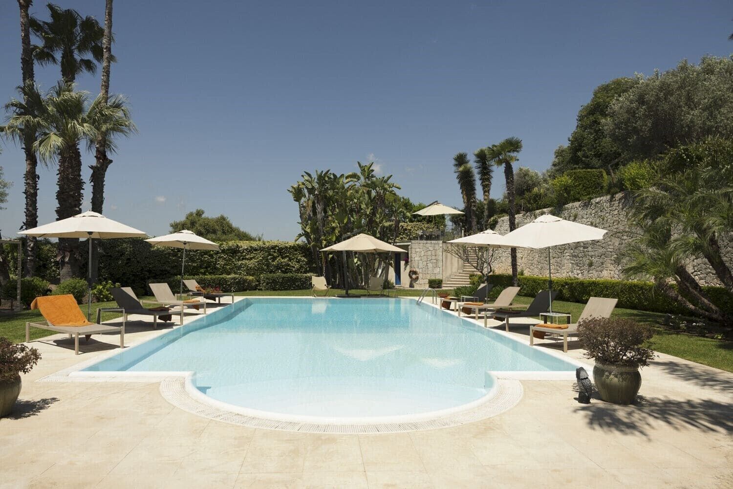 Una piscina rectangular con sillas de descanso y sombrillas, rodeada de palmeras y un muro de piedra.