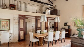 A dining area with a wooden table and white chairs, a staircase leading to a balcony, and a view of the kitchen. - Villa San Tommaso - Four Bedroom Villa, Sleeps 8 (Syracuse)