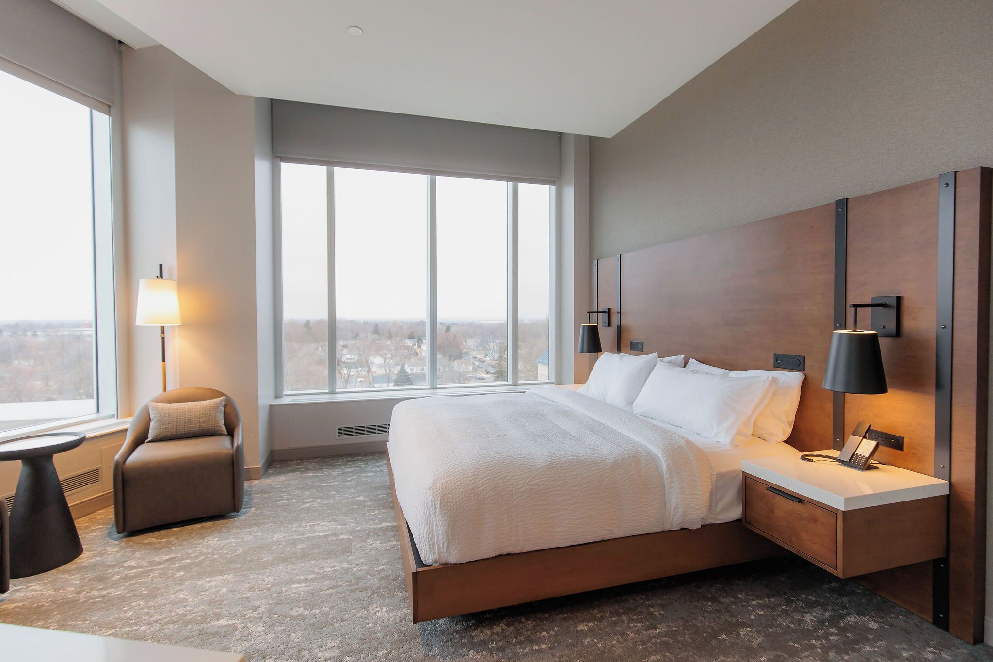 A modern hotel room with a large bed, a desk, a chair, and a large window offering a city view.