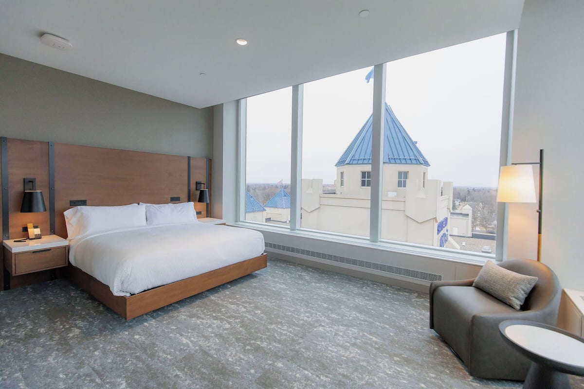 A modern hotel room with a large bed, a nightstand, a bedside table, a chair, a small table, and a large window offering a view of buildings.