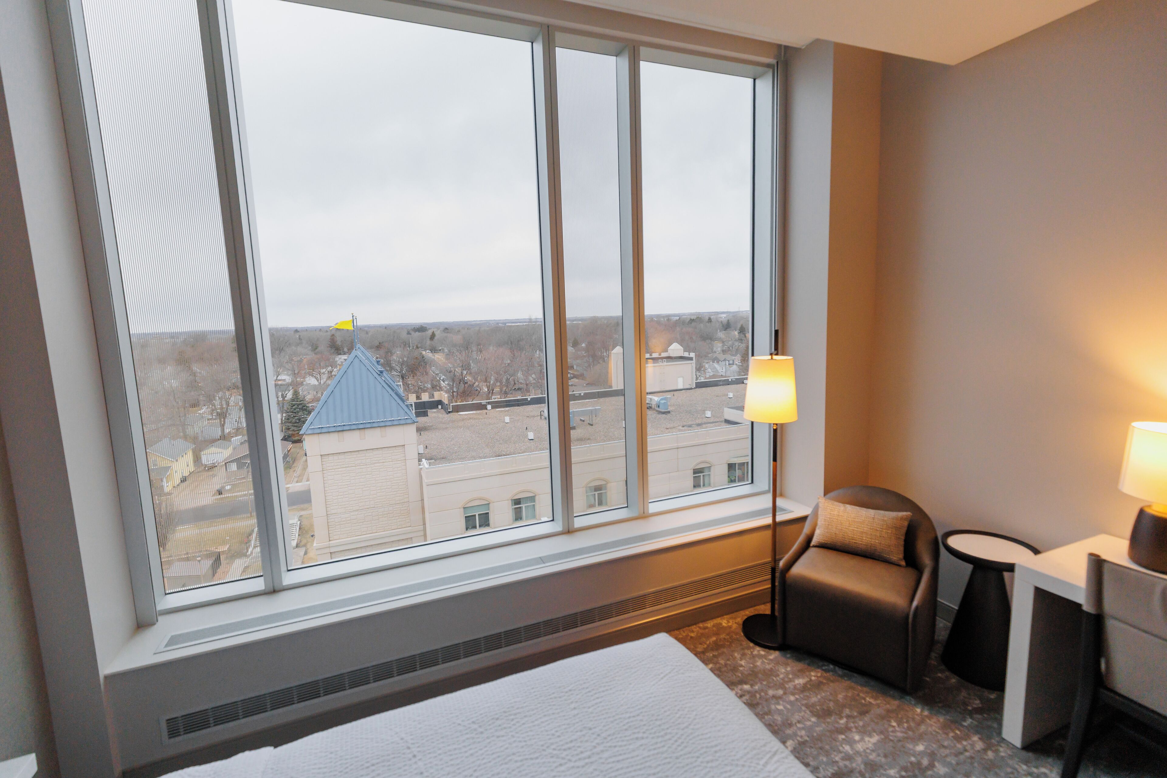A hotel room with a large window, a bed, a chair, a desk, and a lamp.