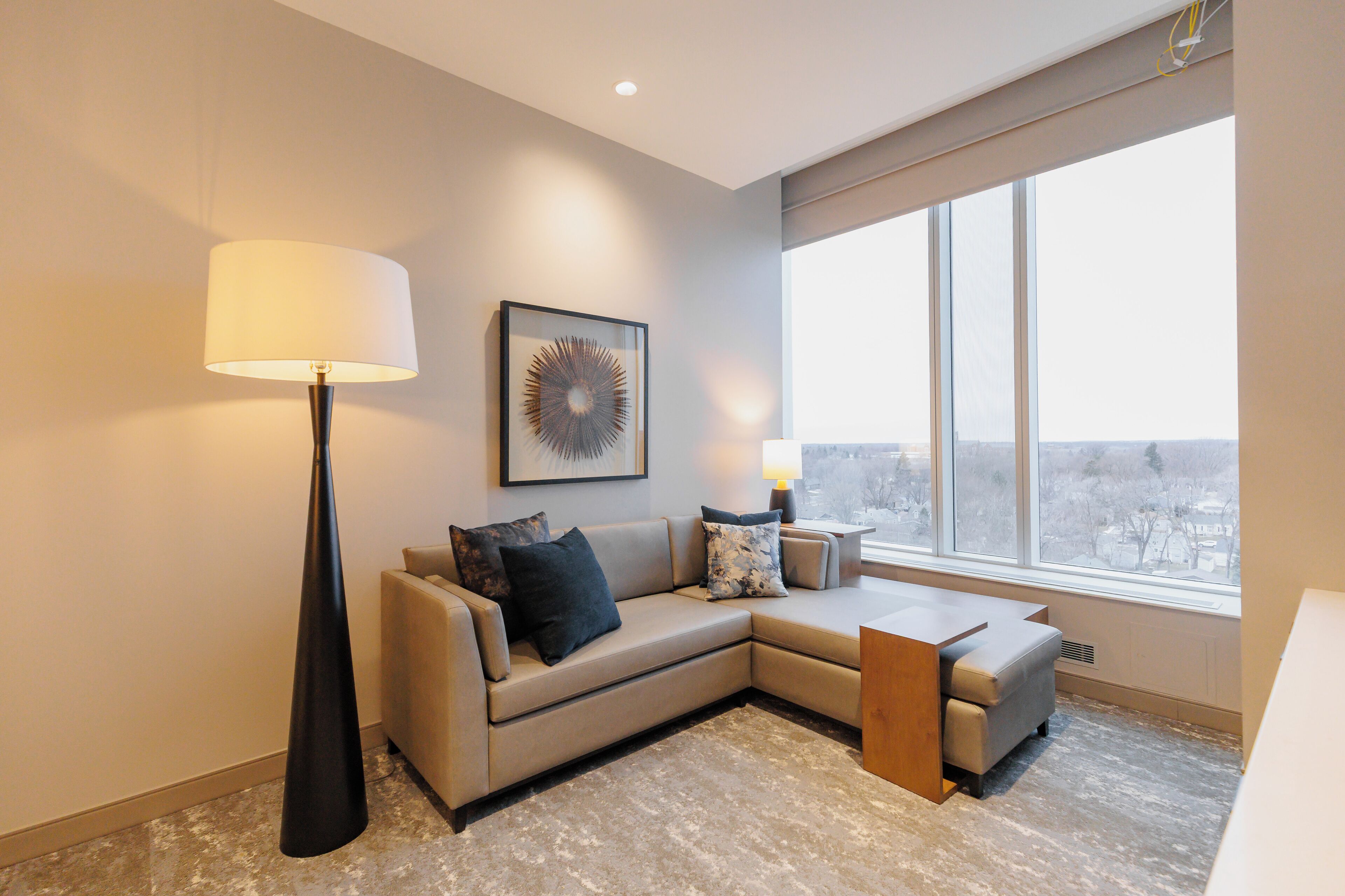 A modern living room with a beige sectional sofa, a floor lamp, a side table with a lamp, and a large window offering a city view.