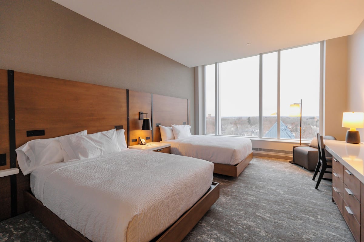 A hotel room with two beds, a desk, and a large window offering a city view.