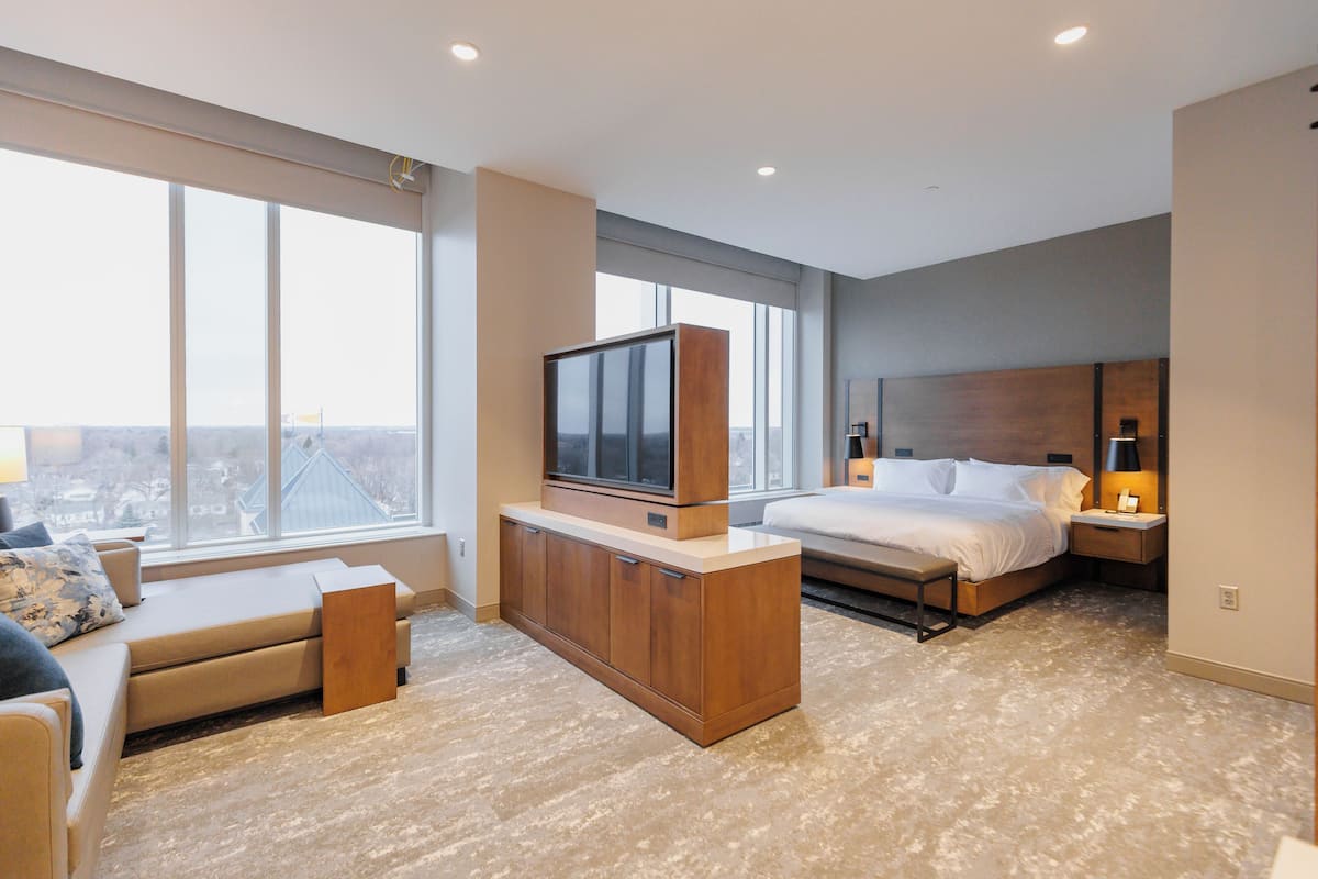 A modern hotel room with a large window, a bed, a TV on a wooden cabinet, a bedside table with a lamp, and a sofa with a bench.