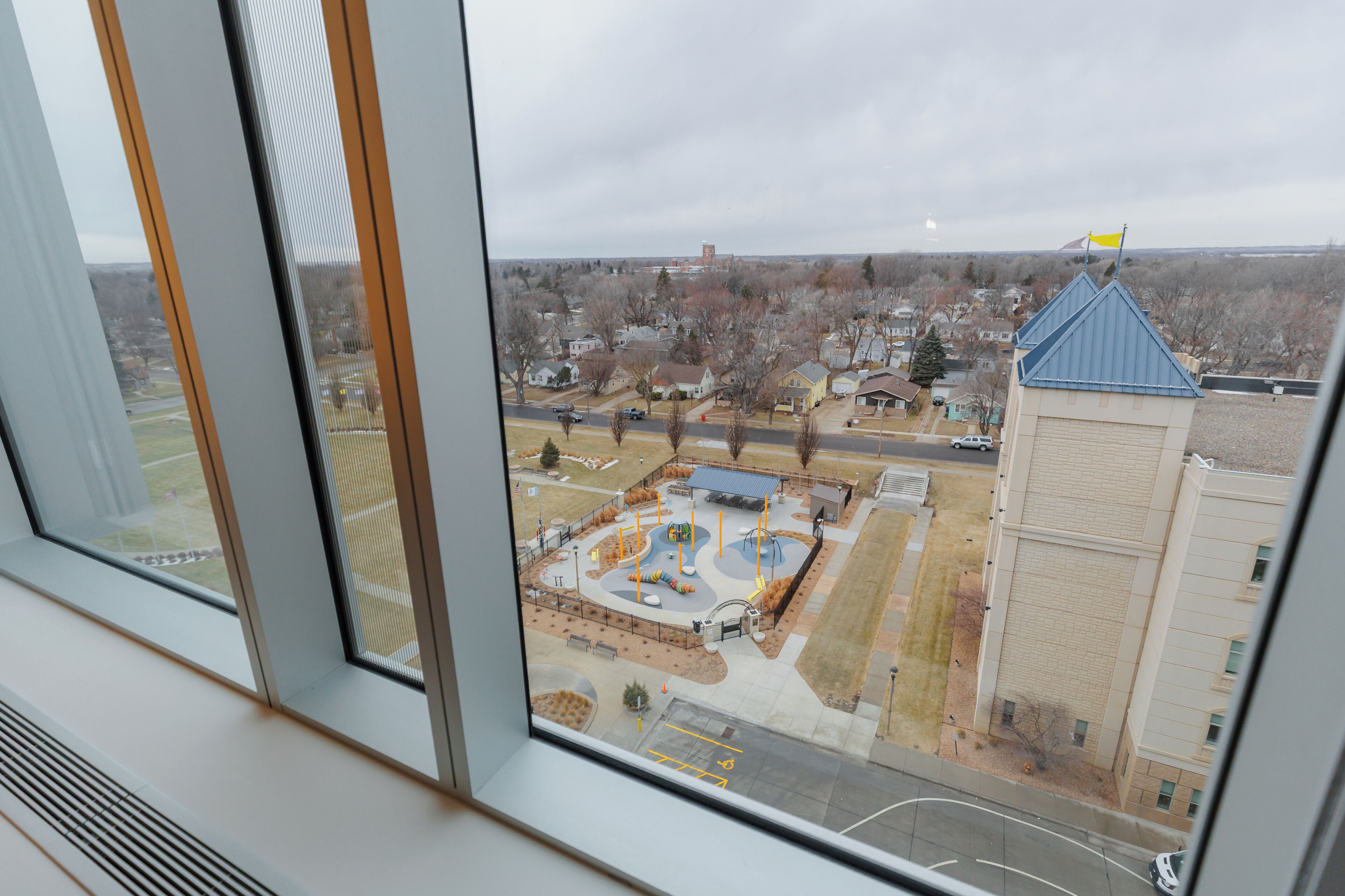 A view through a window of a building with a parking area, a playground, and a distant tower.