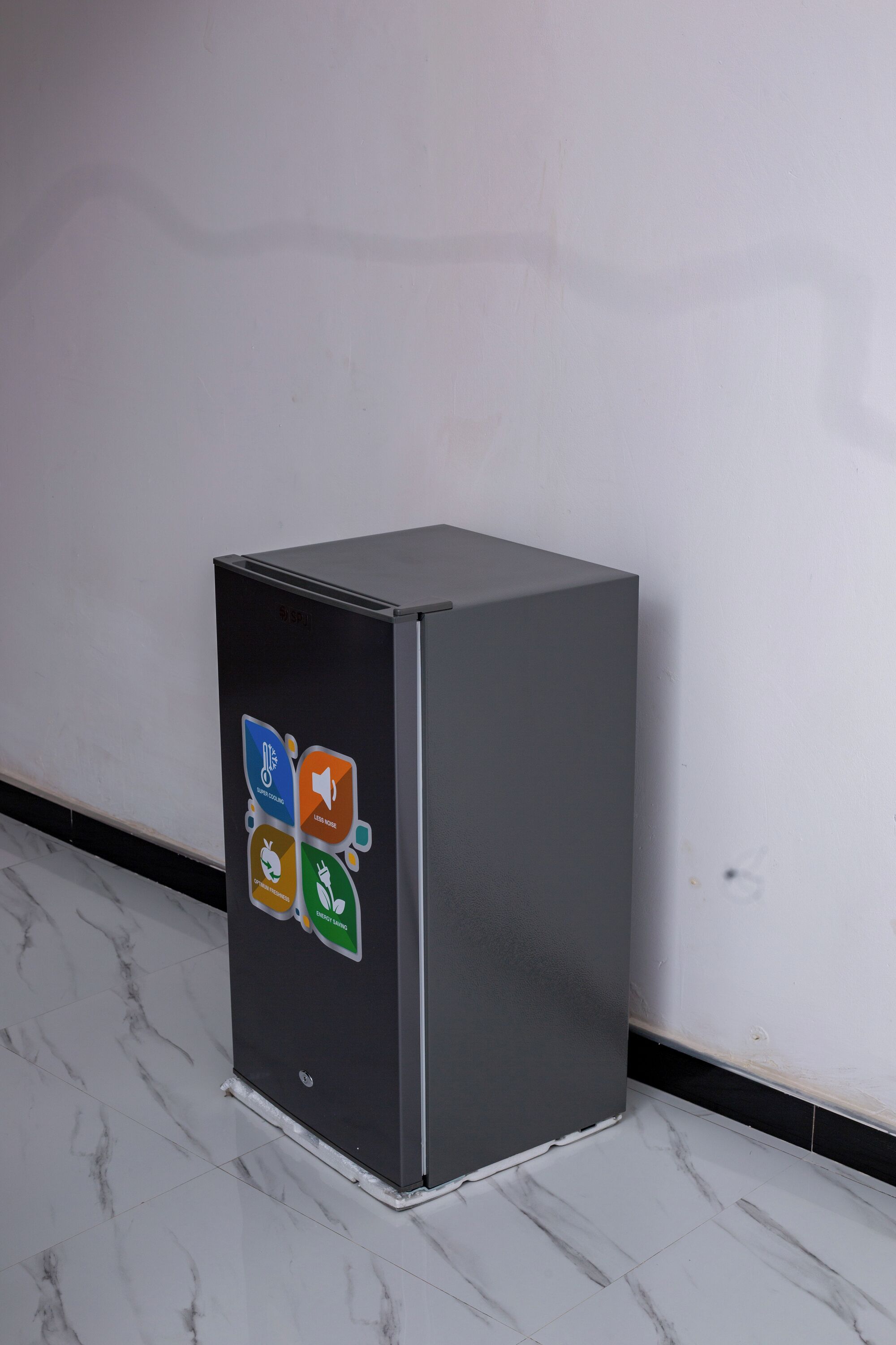 Un refrigerador pequeño y de color oscuro con una pantalla digital en la parte frontal, sobre un piso de mármol y frente a una pared lisa.