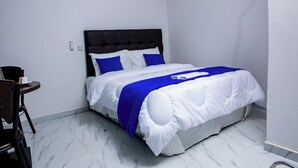Una cama bien hecha con ropa de cama blanca y azul, un cabecero oscuro y una mesita de noche con una taza y un plato.