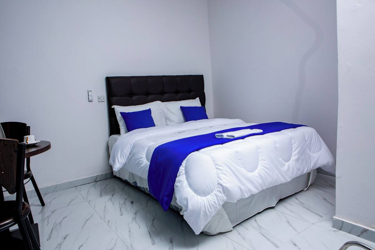 A neatly made bed with white and blue bedding, a dark headboard, and a small table with a cup and saucer beside it.