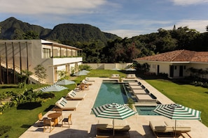 Zona de piscina con sillones de descanso y sombrillas, rodeada por un césped cuidado y con una montaña al fondo.