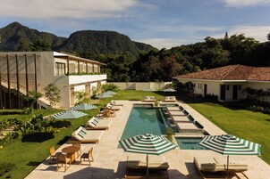 Zona de piscina con sillones de descanso y sombrillas, rodeada por un césped cuidado y con una montaña al fondo.