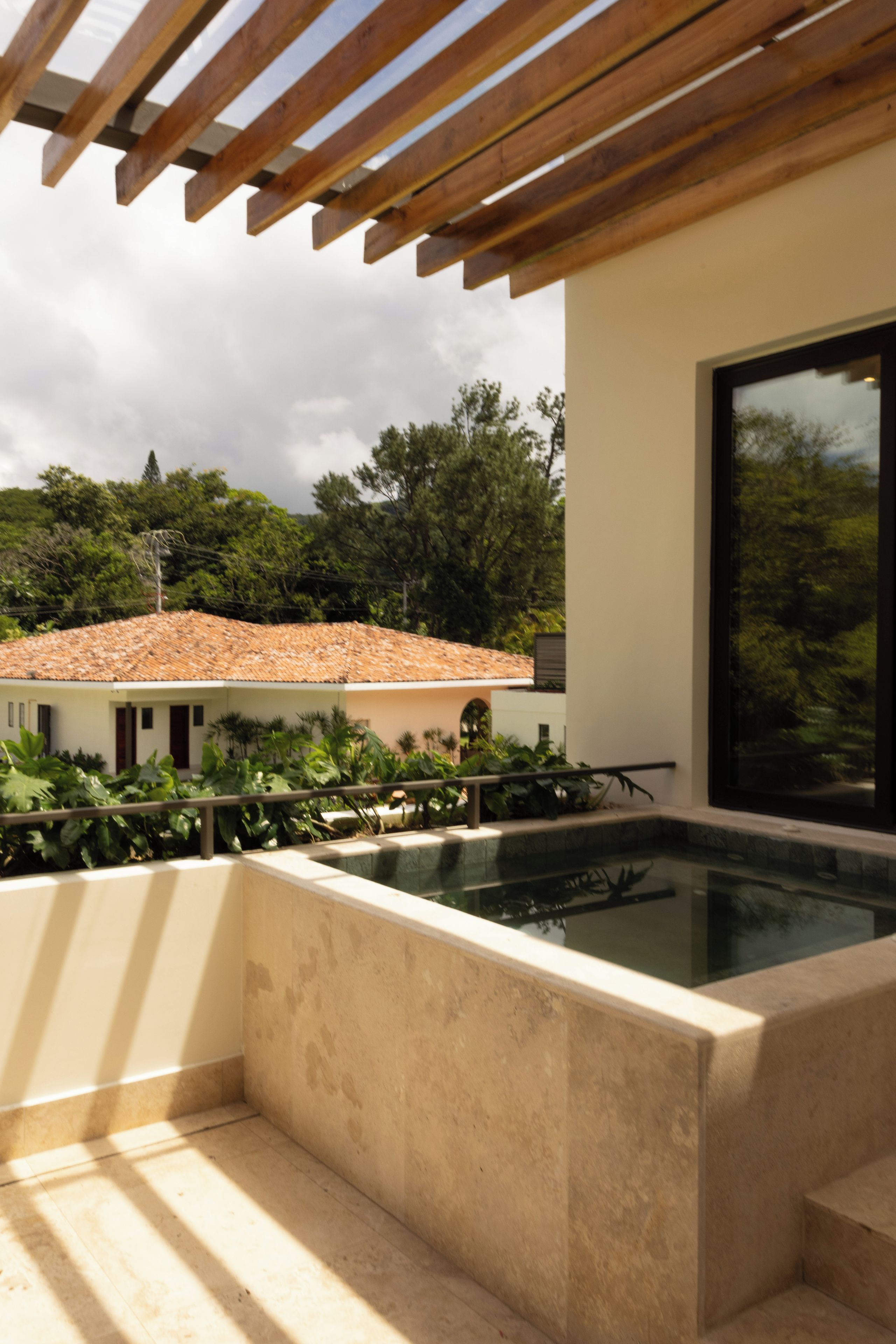 Un espace piscine extérieur moderne, agrémenté d’une pergola, entouré d’une végétation luxuriante et offrant une vue sur un bâtiment traditionnel doté d’un toit en tuiles.