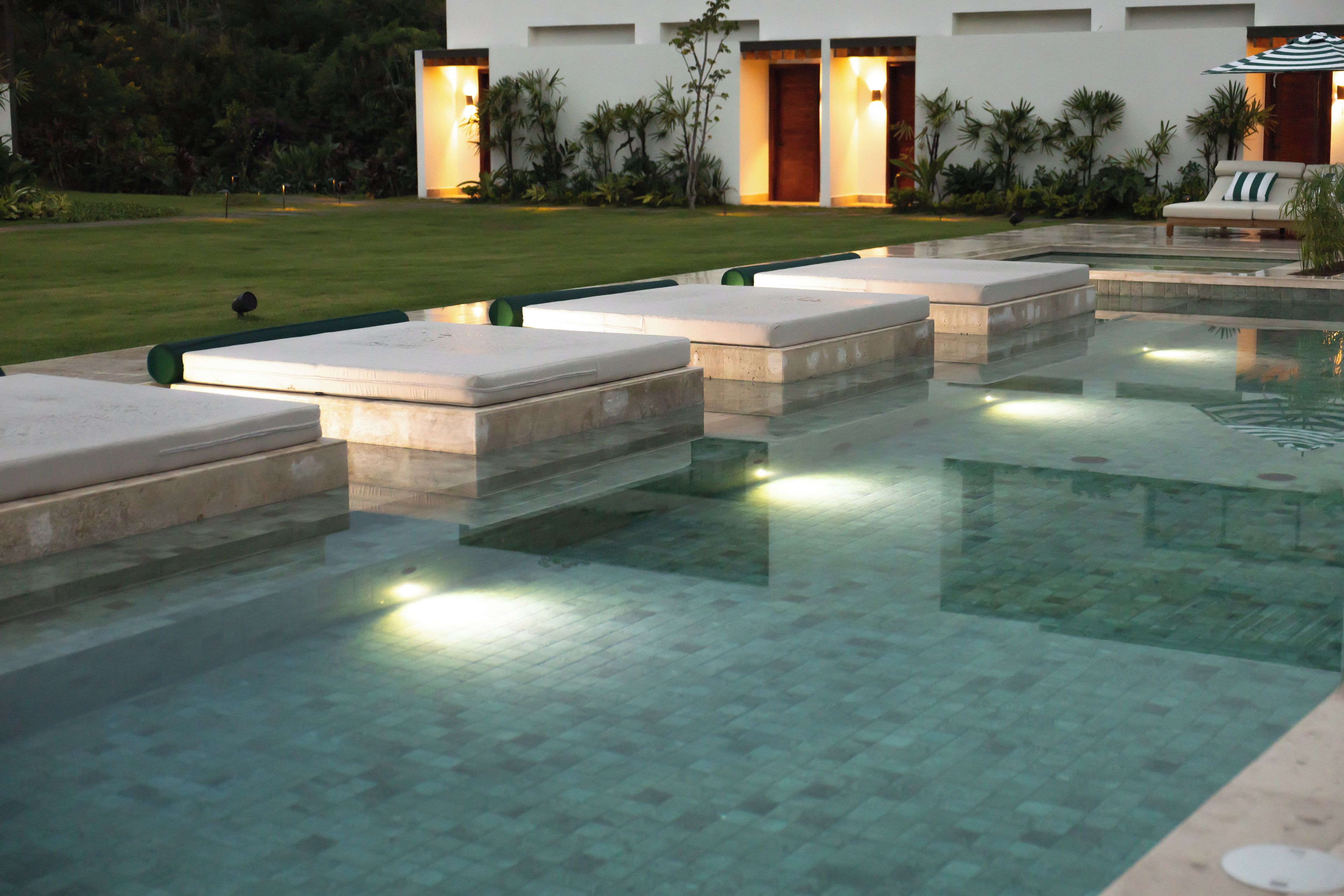 A swimming pool with steps leading into the water, surrounded by a well-maintained lawn and a building with illuminated entryways.