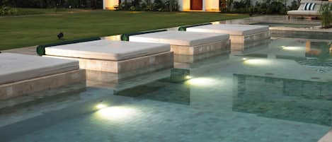 Una piscina con escalones de acceso al agua, rodeada por un césped cuidado y un edificio con entradas iluminadas.